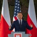 A man in a dark navy suit is speaking at a podium bearing the coat of arms of Poland (white eagle on red background). The setting features two Polish flags flanking the speaker and a United States flag centered behind him, against a military camouflage netting backdrop. The formal arrangement suggests an official bilateral or diplomatic/military event.