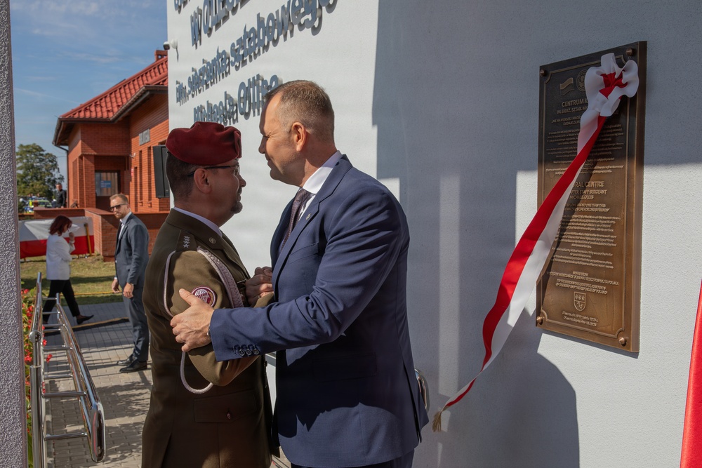 A formal ceremony is taking place outdoors, likely a dedication or inauguration event. A civilian in a navy blue suit appears to be pinning a medal or decoration onto a military officer wearing a maroon beret and olive uniform. A bronze commemorative plaque adorned with a red-and-white ribbon is mounted on a white wall to the right.