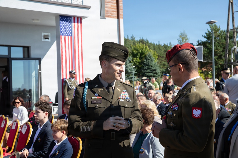 A formal military ceremony is taking place outdoors, featuring both American and Polish military personnel in dress uniforms. Two officers are prominently conversing in the foreground — a U.S. Army officer identified by his name tape as 'WELSH' and a Polish Army officer wearing a red beret with the Polish eagle insignia. An American flag is displayed on the building behind them, and a crowd of seated civilians and military personnel attend the event.