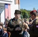 A formal military ceremony is taking place outdoors, featuring both American and Polish military personnel in dress uniforms. Two officers are prominently conversing in the foreground — a U.S. Army officer identified by his name tape as 'WELSH' and a Polish Army officer wearing a red beret with the Polish eagle insignia. An American flag is displayed on the building behind them, and a crowd of seated civilians and military personnel attend the event.
