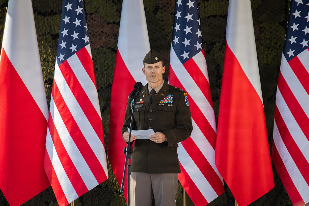 A U.S. Army officer in dress uniform is delivering a speech at a podium microphone. The backdrop consists of alternating American and Polish flags, suggesting a bilateral military or diplomatic ceremony. The officer is holding papers and appears to be reading from prepared remarks.