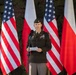 A U.S. Army officer in dress uniform is delivering a speech at a podium microphone. The backdrop consists of alternating American and Polish flags, suggesting a bilateral military or diplomatic ceremony. The officer is holding papers and appears to be reading from prepared remarks.