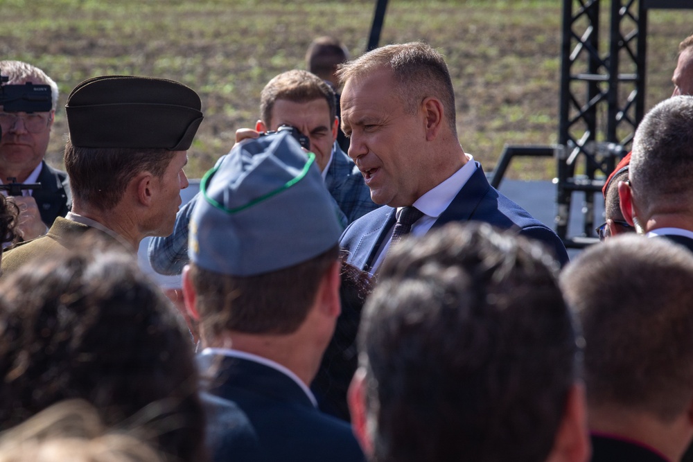 An outdoor gathering or ceremony featuring a mix of civilians and military personnel. A prominent man in a blue suit and dark tie appears to be engaged in conversation with uniformed individuals. The setting includes a stage or platform structure visible in the background.