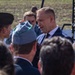An outdoor gathering or ceremony featuring a mix of civilians and military personnel. A prominent man in a blue suit and dark tie appears to be engaged in conversation with uniformed individuals. The setting includes a stage or platform structure visible in the background.