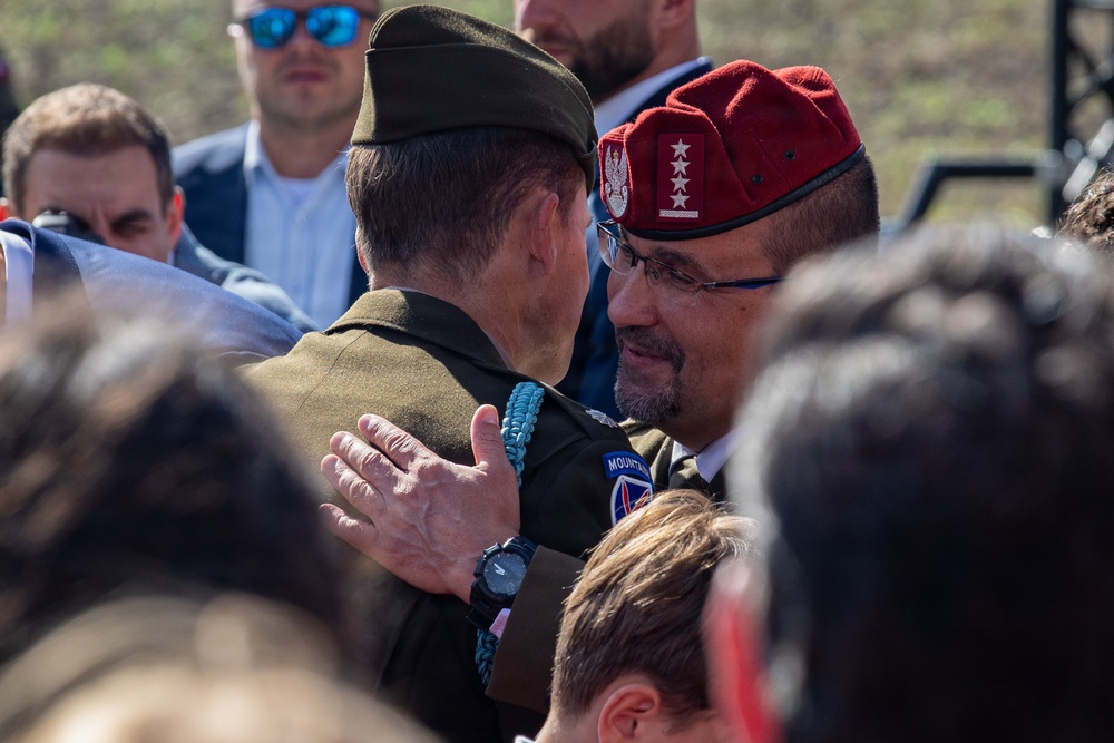 Two military officers are engaged in a greeting or embrace at what appears to be a formal military ceremony or event. One officer wears a US Army uniform with a 10th Mountain Division patch and an olive garrison cap, while the other wears a red beret with a Polish eagle emblem and three stars, suggesting a senior Polish military officer. The scene is surrounded by other attendees in both military and civilian attire.