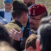 Two military officers are engaged in a greeting or embrace at what appears to be a formal military ceremony or event. One officer wears a US Army uniform with a 10th Mountain Division patch and an olive garrison cap, while the other wears a red beret with a Polish eagle emblem and three stars, suggesting a senior Polish military officer. The scene is surrounded by other attendees in both military and civilian attire.