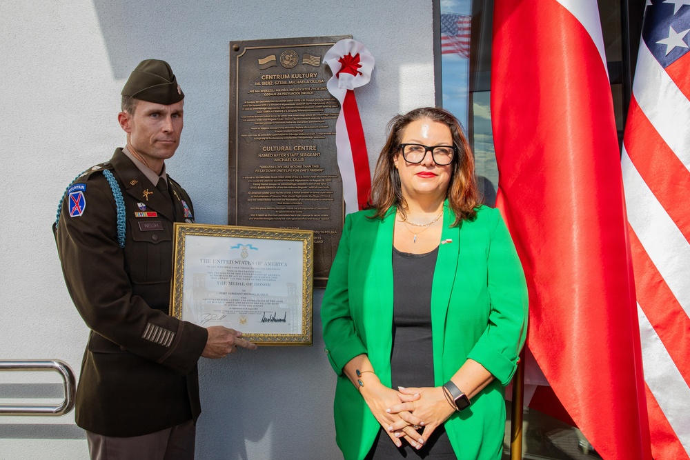 A U.S. Army officer and a civilian woman stand before a commemorative bronze plaque on a building wall. The officer holds a framed Medal of Honor citation from the United States of America. The plaque identifies a Cultural Centre named after Staff Sergeant Michael Ollis, with flags of Poland and the United States visible in the background.
