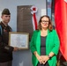 A U.S. Army officer and a civilian woman stand before a commemorative bronze plaque on a building wall. The officer holds a framed Medal of Honor citation from the United States of America. The plaque identifies a Cultural Centre named after Staff Sergeant Michael Ollis, with flags of Poland and the United States visible in the background.