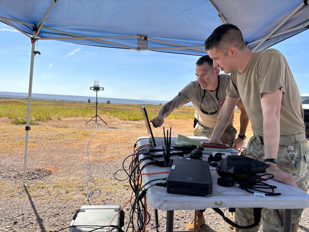 Two military personnel in tan/OCP uniforms are working at a field operations station set up under a portable canopy tent. They are examining a laptop computer surrounded by networking equipment, cables, and communication devices on a folding table. In the background, an antenna or sensor array is mounted on a tripod in an open field environment.