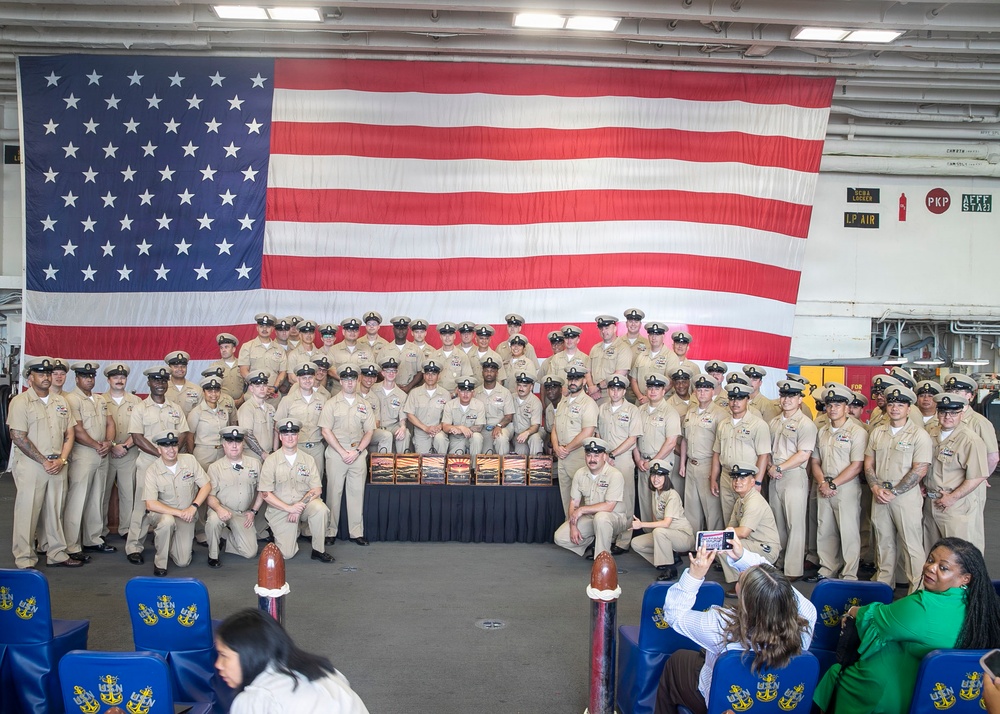 A large group of U.S. Navy personnel in khaki service uniforms pose for a formal group photograph in front of an oversized American flag. The setting appears to be a ship's hangar bay, likely aboard a naval vessel, during what appears to be a chief petty officer pinning or advancement ceremony. Civilian guests and family members are seated in the foreground watching the event.