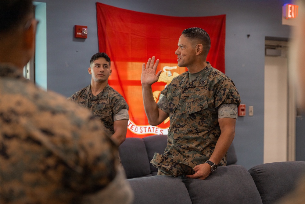 Two U.S. Marines in digital camouflage uniforms are seated in what appears to be a lounge or common area. The Marine in the foreground is raising his right hand, possibly administering or taking an oath, while another Marine observes. A red United States Marine Corps flag is displayed on the wall behind them.