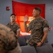 Two U.S. Marines in digital camouflage uniforms are seated in what appears to be a lounge or common area. The Marine in the foreground is raising his right hand, possibly administering or taking an oath, while another Marine observes. A red United States Marine Corps flag is displayed on the wall behind them.