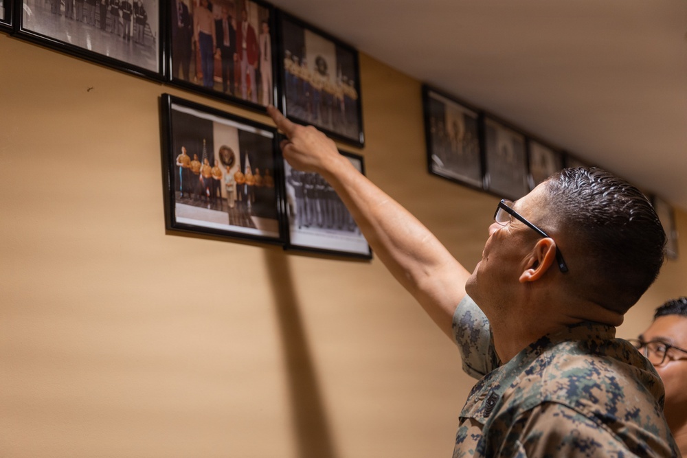 A person in U.S. Marine Corps camouflage uniform is pointing at one of several framed photographs mounted on a beige wall. The photos appear to be historical or commemorative military group photographs arranged in a row. A second individual wearing glasses is partially visible behind the primary subject.