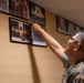 A person in U.S. Marine Corps camouflage uniform is pointing at one of several framed photographs mounted on a beige wall. The photos appear to be historical or commemorative military group photographs arranged in a row. A second individual wearing glasses is partially visible behind the primary subject.