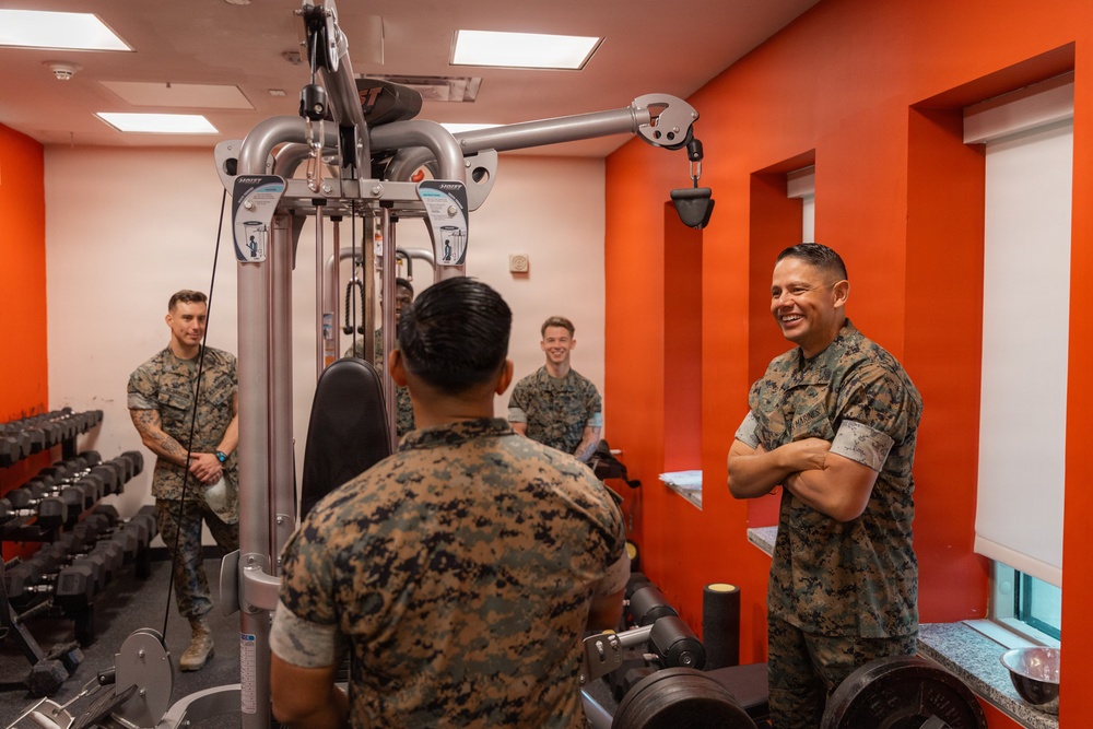 A group of U.S. Marines in camouflage uniforms are gathered inside a fitness facility with bright orange walls. A senior Marine on the right, arms crossed and smiling, appears to be engaged in conversation with the others. The setting suggests an informal interaction or inspection of a gym facility.