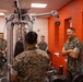A group of U.S. Marines in camouflage uniforms are gathered inside a fitness facility with bright orange walls. A senior Marine on the right, arms crossed and smiling, appears to be engaged in conversation with the others. The setting suggests an informal interaction or inspection of a gym facility.