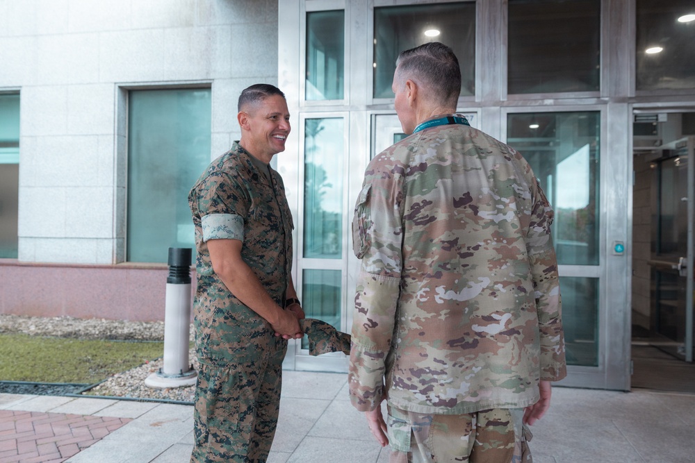 Two military personnel in different camouflage uniforms are shaking hands outside a modern building. The individual on the left is wearing USMC MARPAT (Marine Pattern) digital camouflage and is smiling, while the individual on the right is wearing US Army/Air Force OCP (Operational Camouflage Pattern) and has their back to the camera. The setting appears to be an official military installation or headquarters building.