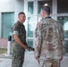 Two military personnel in different camouflage uniforms are shaking hands outside a modern building. The individual on the left is wearing USMC MARPAT (Marine Pattern) digital camouflage and is smiling, while the individual on the right is wearing US Army/Air Force OCP (Operational Camouflage Pattern) and has their back to the camera. The setting appears to be an official military installation or headquarters building.