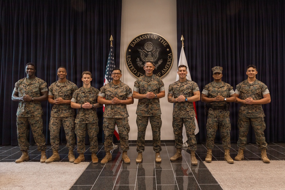 Eight U.S. Marines in MARPAT camouflage uniforms pose for a formal group photograph inside a U.S. Embassy. They are standing in front of the official Embassy of the United States of America seal, flanked by the American flag and what appears to be a host nation flag. The setting is an official diplomatic facility lobby with dark curtain backdrop.