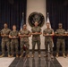 Eight U.S. Marines in MARPAT camouflage uniforms pose for a formal group photograph inside a U.S. Embassy. They are standing in front of the official Embassy of the United States of America seal, flanked by the American flag and what appears to be a host nation flag. The setting is an official diplomatic facility lobby with dark curtain backdrop.