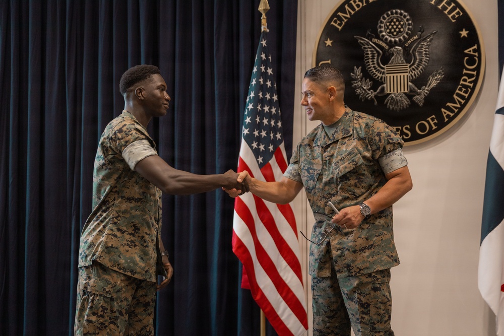 Two U.S. Marines in MARPAT digital camouflage uniforms are shaking hands in front of an American flag and the official seal of the Embassy of the United States of America. The setting appears to be a formal military ceremony or recognition event held at a U.S. Embassy. The right individual has 'U.S. MARINES' visible on their uniform and is smiling.