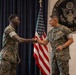 Two U.S. Marines in MARPAT digital camouflage uniforms are shaking hands in front of an American flag and the official seal of the Embassy of the United States of America. The setting appears to be a formal military ceremony or recognition event held at a U.S. Embassy. The right individual has 'U.S. MARINES' visible on their uniform and is smiling.