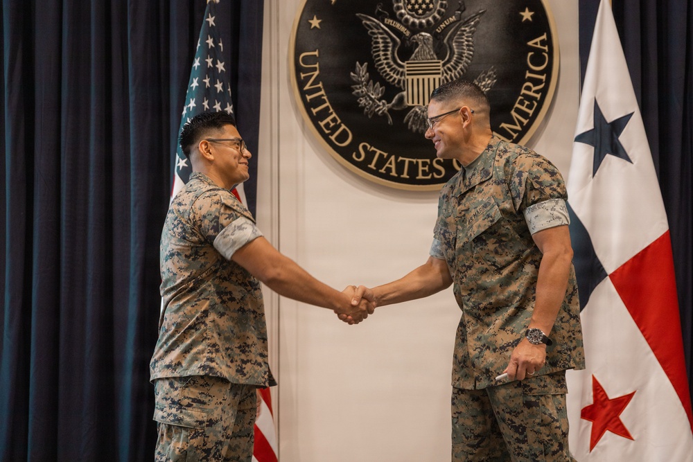 Two U.S. Marine Corps personnel in MARPAT digital camouflage uniforms are shaking hands in front of an official backdrop featuring the Great Seal of the United States of America. The setting appears to be a formal military ceremony, likely a promotion, award, or recognition event. The flags of the United States and Panama are visible in the background, suggesting this event took place at a U.S. diplomatic or military facility in Panama.
