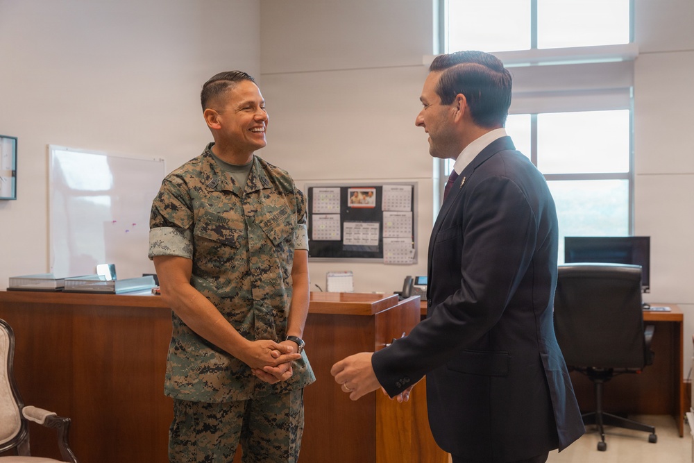 Two individuals are engaged in a friendly conversation inside what appears to be a military office setting. One person is wearing U.S. Marine Corps MARPAT digital camouflage uniform, while the other is dressed in a dark business suit. The interaction appears cordial and informal, with the uniformed individual smiling broadly.