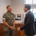 Two individuals are engaged in a friendly conversation inside what appears to be a military office setting. One person is wearing U.S. Marine Corps MARPAT digital camouflage uniform, while the other is dressed in a dark business suit. The interaction appears cordial and informal, with the uniformed individual smiling broadly.