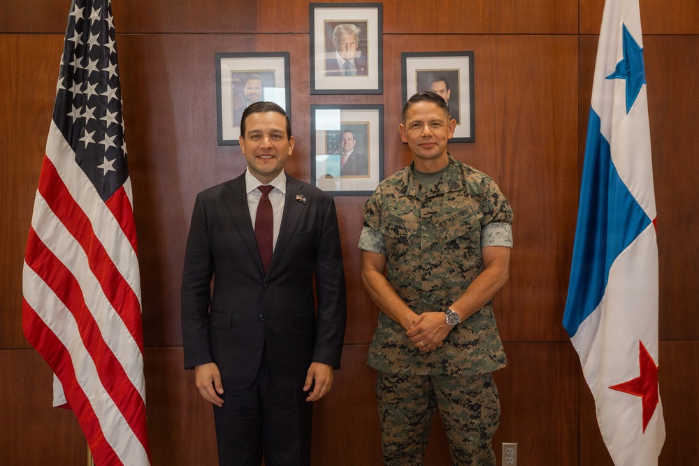 Two individuals pose for a formal photograph in what appears to be an official government or diplomatic office setting. One person is wearing a dark business suit with a red tie and a small lapel pin, while the other is dressed in U.S. Marine Corps digital camouflage (MARPAT) uniform. The setting features both an American flag and a Panamanian flag, suggesting a bilateral diplomatic or military meeting.
