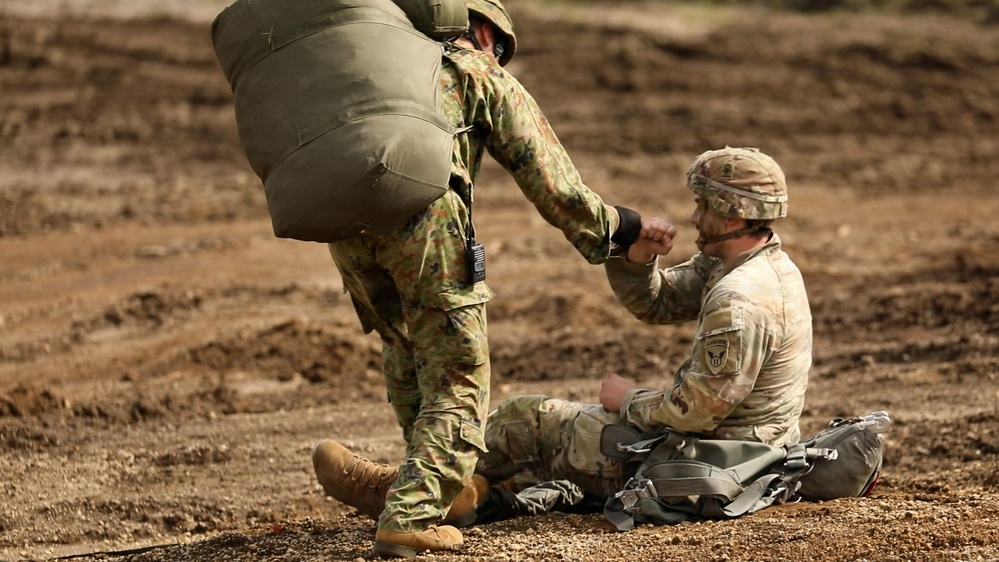 Two military personnel are visible on a dirt terrain, with one soldier seated on the ground and another standing over them extending a hand, appearing to assist them up. The standing soldier carries a large bundled pack on their back, consistent with parachute or airborne equipment. The scene appears to depict a post-landing assist during an airborne or parachute training exercise.