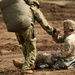 Two military personnel are visible on a dirt terrain, with one soldier seated on the ground and another standing over them extending a hand, appearing to assist them up. The standing soldier carries a large bundled pack on their back, consistent with parachute or airborne equipment. The scene appears to depict a post-landing assist during an airborne or parachute training exercise.
