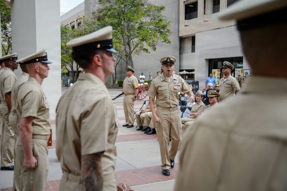 A U.S. Navy military ceremony or formation is taking place in an outdoor urban plaza. A central figure, appearing to be a senior enlisted sailor or officer, is speaking or issuing commands while walking between rows of uniformed personnel. Spectators are seated in the background observing the event.