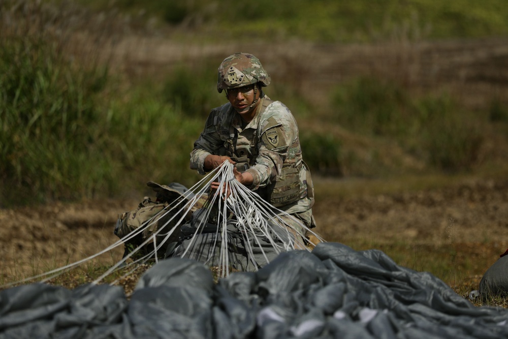 A U.S. Army soldier in full combat uniform and helmet is gathering and bundling parachute suspension lines after a jump landing. The soldier is kneeling on open ground, collecting the white nylon cords connected to a dark-colored parachute canopy. This appears to be a post-jump recovery operation on a drop zone.