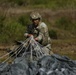 A U.S. Army soldier in full combat uniform and helmet is gathering and bundling parachute suspension lines after a jump landing. The soldier is kneeling on open ground, collecting the white nylon cords connected to a dark-colored parachute canopy. This appears to be a post-jump recovery operation on a drop zone.