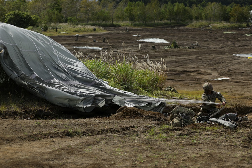 A military paratrooper in camouflage uniform is on the ground gathering and recovering a large gray parachute after a landing on a dirt drop zone. The scene appears to be a military airborne exercise with multiple personnel and parachutes visible across the field in the background. The landing zone is a wide open area of disturbed earth with sparse vegetation.