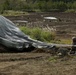A military paratrooper in camouflage uniform is on the ground gathering and recovering a large gray parachute after a landing on a dirt drop zone. The scene appears to be a military airborne exercise with multiple personnel and parachutes visible across the field in the background. The landing zone is a wide open area of disturbed earth with sparse vegetation.