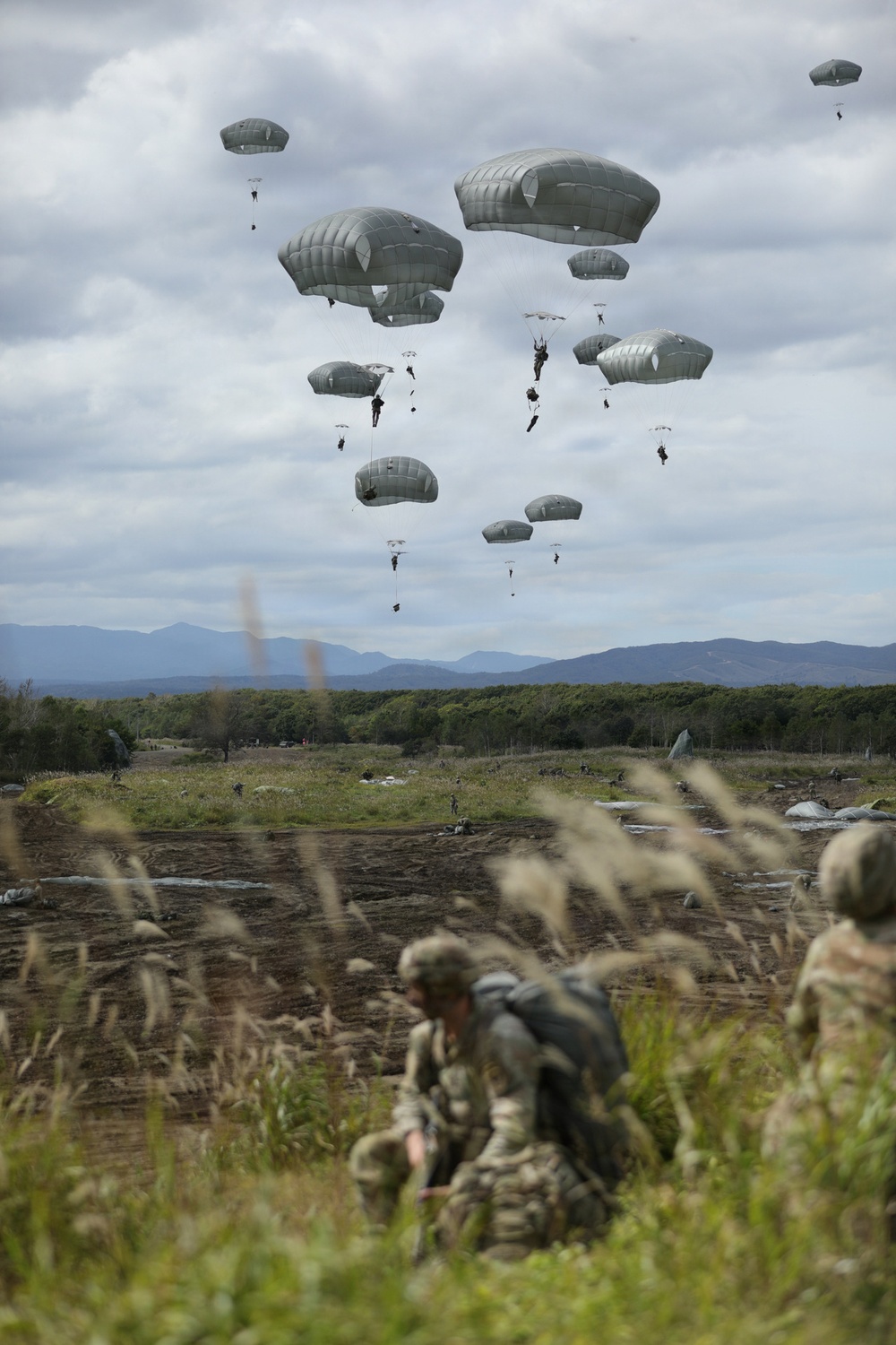 A large-scale military airborne operation showing numerous paratroopers descending under olive-drab/gray parachutes over an open drop zone. Multiple soldiers have already landed on the ground in the foreground, while others are still descending from altitude. The backdrop features a treeline and distant mountain range under overcast skies.
