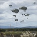 A large-scale military airborne operation showing numerous paratroopers descending under olive-drab/gray parachutes over an open drop zone. Multiple soldiers have already landed on the ground in the foreground, while others are still descending from altitude. The backdrop features a treeline and distant mountain range under overcast skies.
