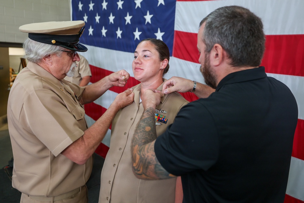 A military promotion or pinning ceremony is taking place in front of a large American flag. A female service member in U.S. Navy tan/khaki uniform is having insignia pinned to her collar by two individuals — an older man in uniform with a cover (hat) and a civilian man in a black shirt. The service member is smiling, suggesting this is a celebratory milestone event.