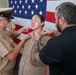 A military promotion or pinning ceremony is taking place in front of a large American flag. A female service member in U.S. Navy tan/khaki uniform is having insignia pinned to her collar by two individuals — an older man in uniform with a cover (hat) and a civilian man in a black shirt. The service member is smiling, suggesting this is a celebratory milestone event.