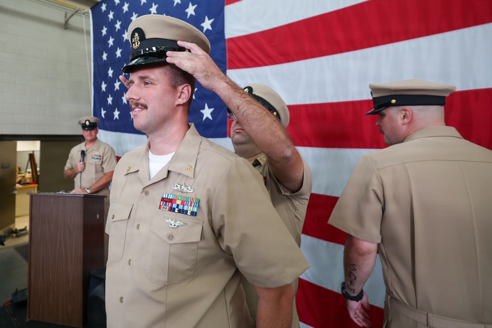 A U.S. Navy promotion or advancement ceremony is taking place in front of a large American flag. A sailor is having a chief petty officer cover (hat) placed on his head by another service member, indicating a pinning or frocking ceremony. A uniformed officer speaks at a podium in the background while other personnel observe.