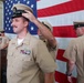 A U.S. Navy promotion or advancement ceremony is taking place in front of a large American flag. A sailor is having a chief petty officer cover (hat) placed on his head by another service member, indicating a pinning or frocking ceremony. A uniformed officer speaks at a podium in the background while other personnel observe.