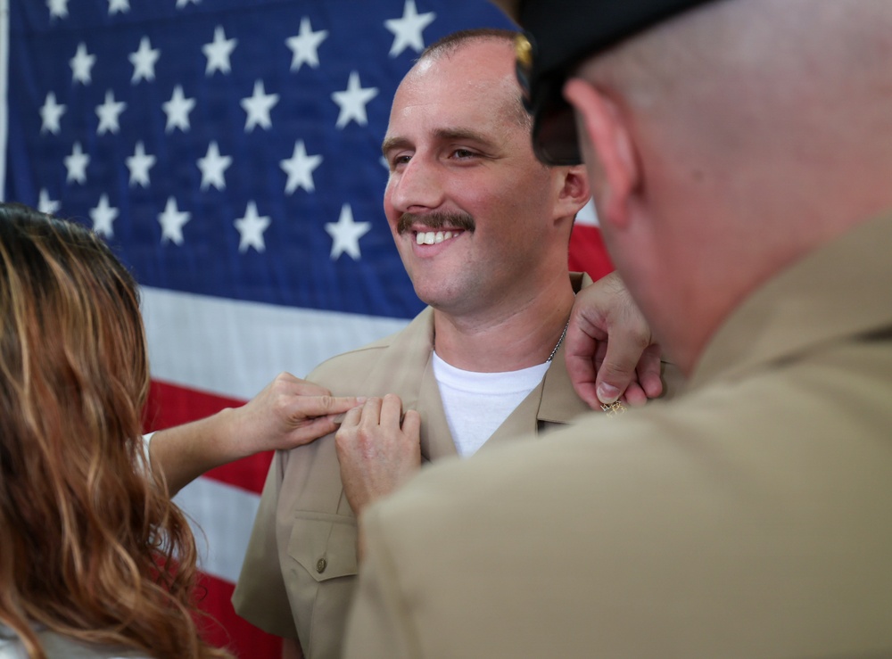 A smiling military serviceman in a tan/khaki uniform is having insignia or rank devices pinned onto his collar by two individuals — a woman on his left and an older man in uniform on his right. An American flag serves as the backdrop, suggesting this is a formal military promotion or pinning ceremony. The subject appears proud and happy during what is clearly a significant milestone event.