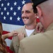 A smiling military serviceman in a tan/khaki uniform is having insignia or rank devices pinned onto his collar by two individuals — a woman on his left and an older man in uniform on his right. An American flag serves as the backdrop, suggesting this is a formal military promotion or pinning ceremony. The subject appears proud and happy during what is clearly a significant milestone event.