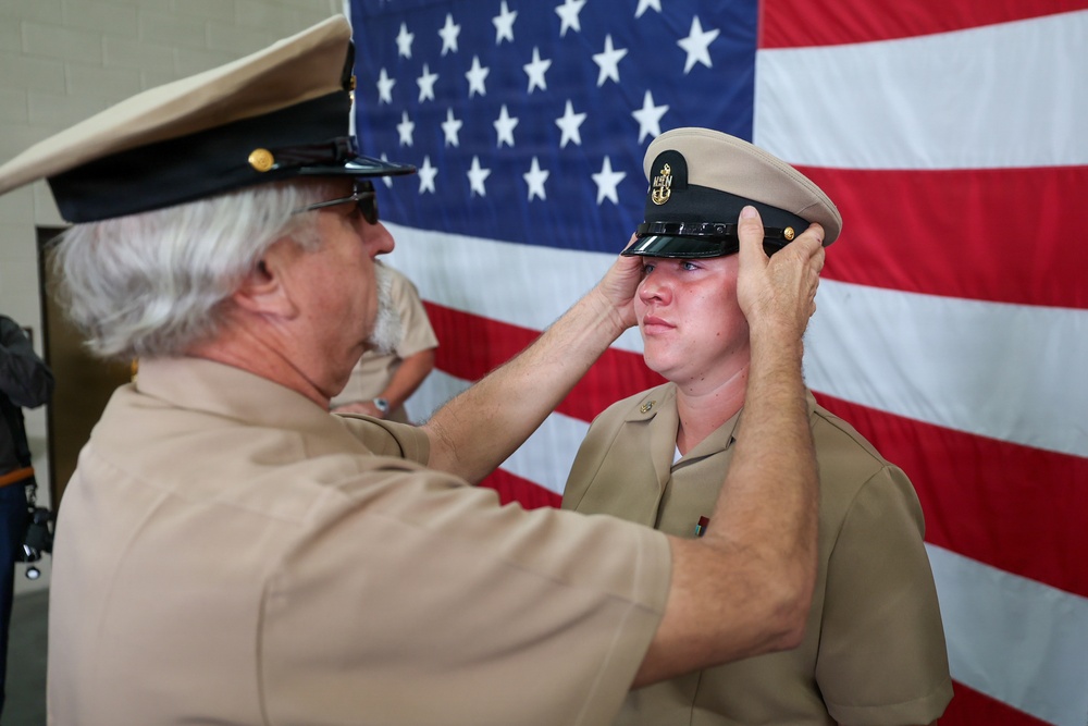 An older military servicemember in U.S. Navy khaki uniform is placing or adjusting a cover (combination cap) on a younger servicemember's head during what appears to be a promotion or pinning ceremony. Both individuals are wearing U.S. Navy Service Khaki uniforms, and a large American flag serves as the backdrop. The moment appears ceremonial and significant, possibly marking a chief petty officer advancement.