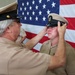 An older military servicemember in U.S. Navy khaki uniform is placing or adjusting a cover (combination cap) on a younger servicemember's head during what appears to be a promotion or pinning ceremony. Both individuals are wearing U.S. Navy Service Khaki uniforms, and a large American flag serves as the backdrop. The moment appears ceremonial and significant, possibly marking a chief petty officer advancement.