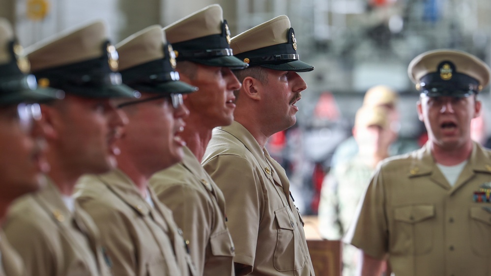A group of U.S. Navy personnel in khaki service uniforms and combination covers (peaked caps) are standing in a formal formation. One individual on the right appears to be speaking or issuing commands, facing the line of sailors. The setting appears to be an indoor military facility, possibly a hangar or large ceremonial hall.