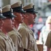 A group of U.S. Navy personnel in khaki service uniforms and combination covers (peaked caps) are standing in a formal formation. One individual on the right appears to be speaking or issuing commands, facing the line of sailors. The setting appears to be an indoor military facility, possibly a hangar or large ceremonial hall.