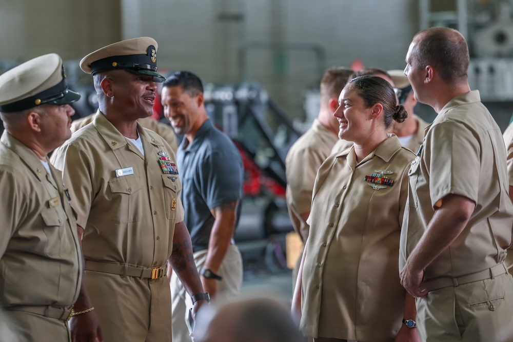 A group of U.S. Navy personnel in tan/khaki service uniforms are gathered in what appears to be a hangar or industrial facility. Two central figures — a senior enlisted male sailor wearing a combination cover and a female sailor — appear to be engaged in a friendly conversation, both smiling. The setting suggests an informal gathering or post-ceremony interaction.